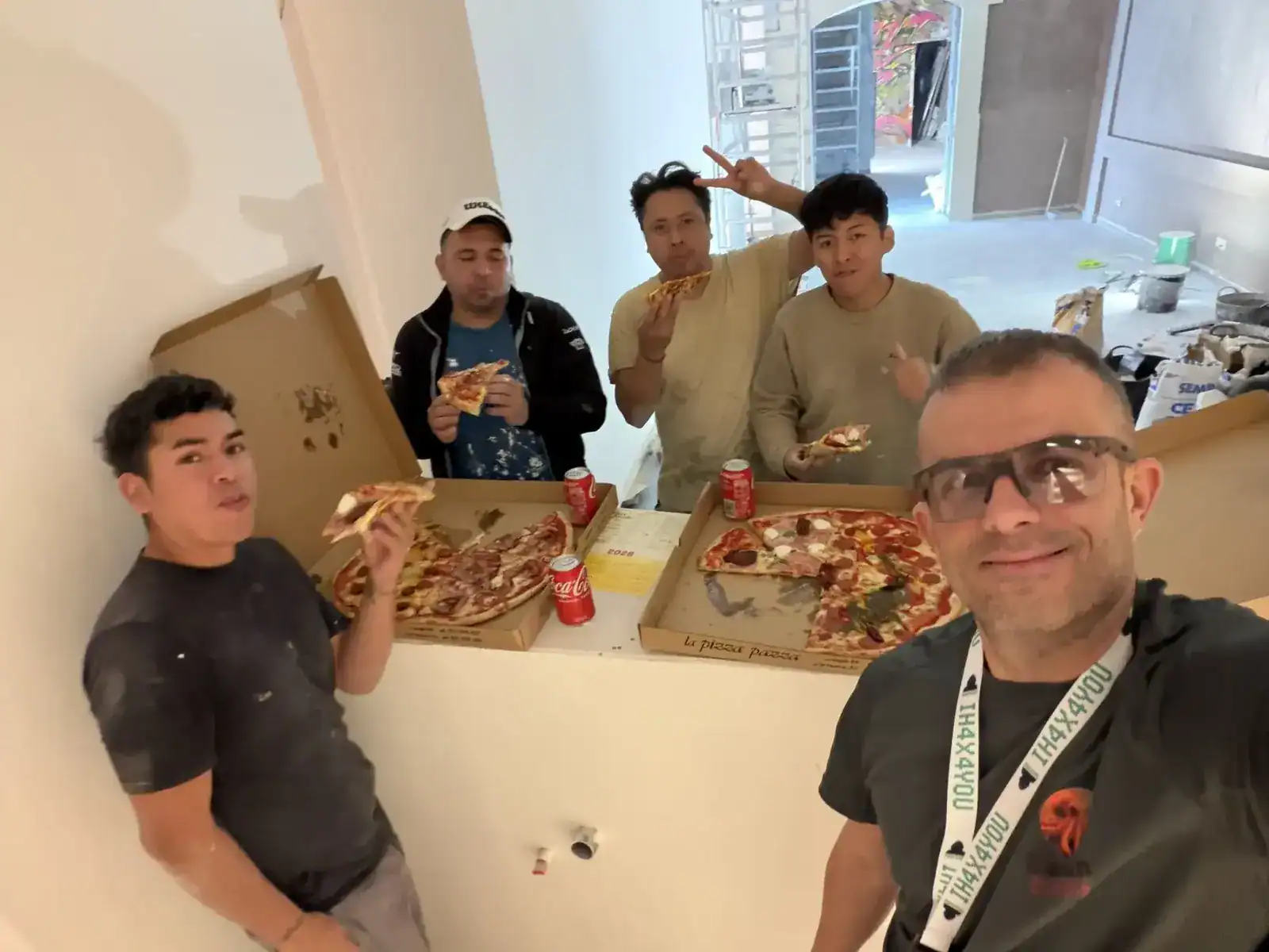Alt: Grupo de amigos comiendo pizza en un espacio en construcción con máquinas y materiales visibles.